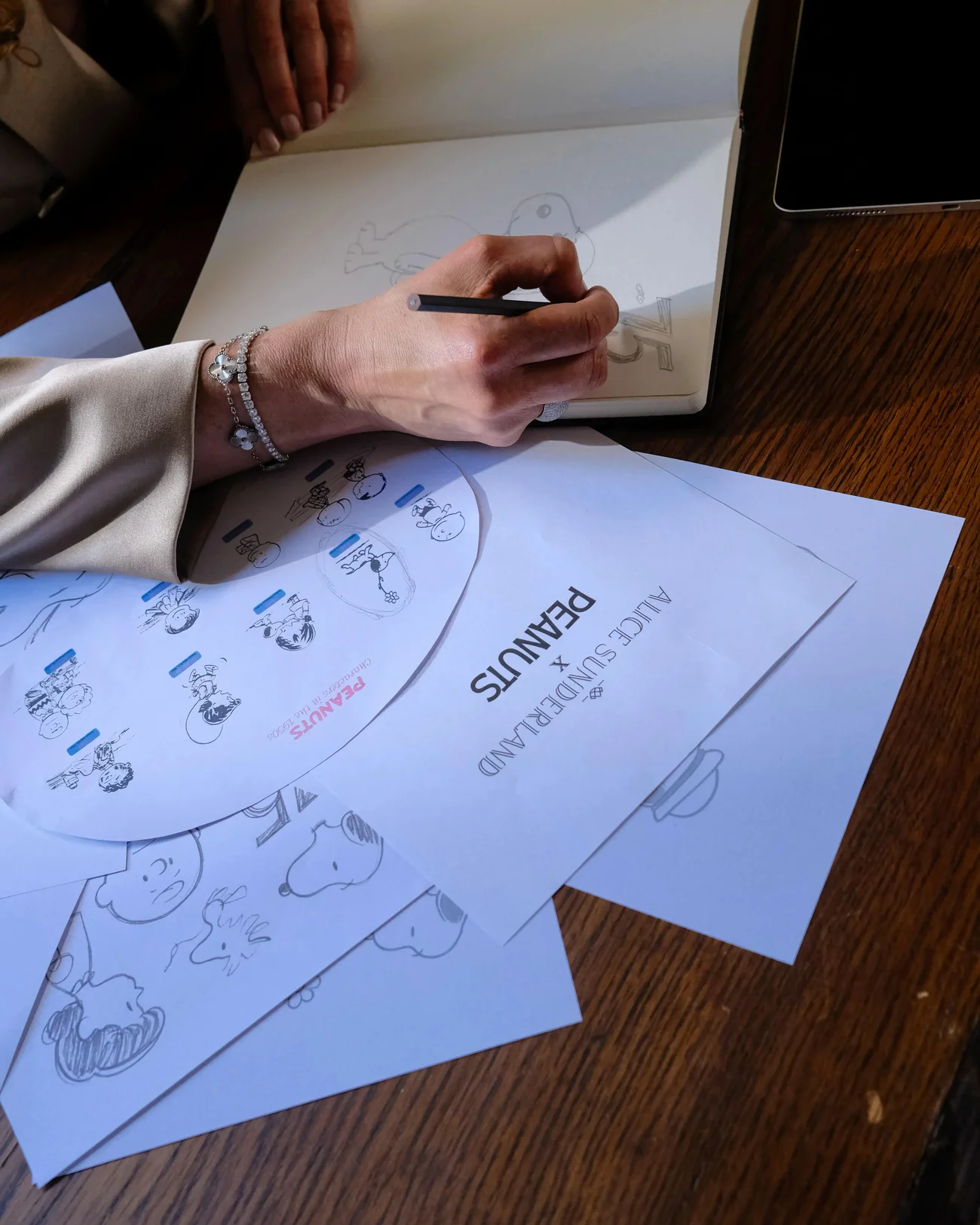 Alice Sunderland sketching Snoopy pendant designs at her desk, diamond bracelets on her wrist, Peanuts character sheets across the wood