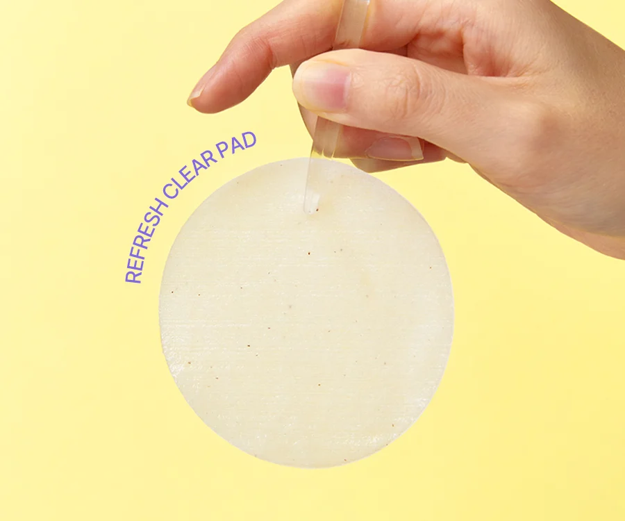 A hand holding the Refresh Clear Pad against a soft yellow backdrop